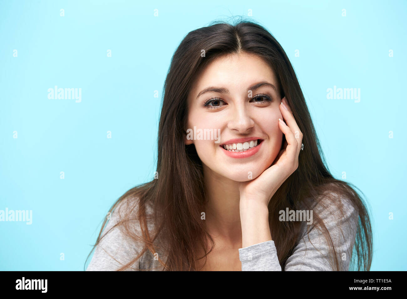 Retrato de una joven y bella mujer caucásica, mirando a la cámara sonriendo, con la mano en la mejilla, aislado sobre fondo azul. Foto de stock