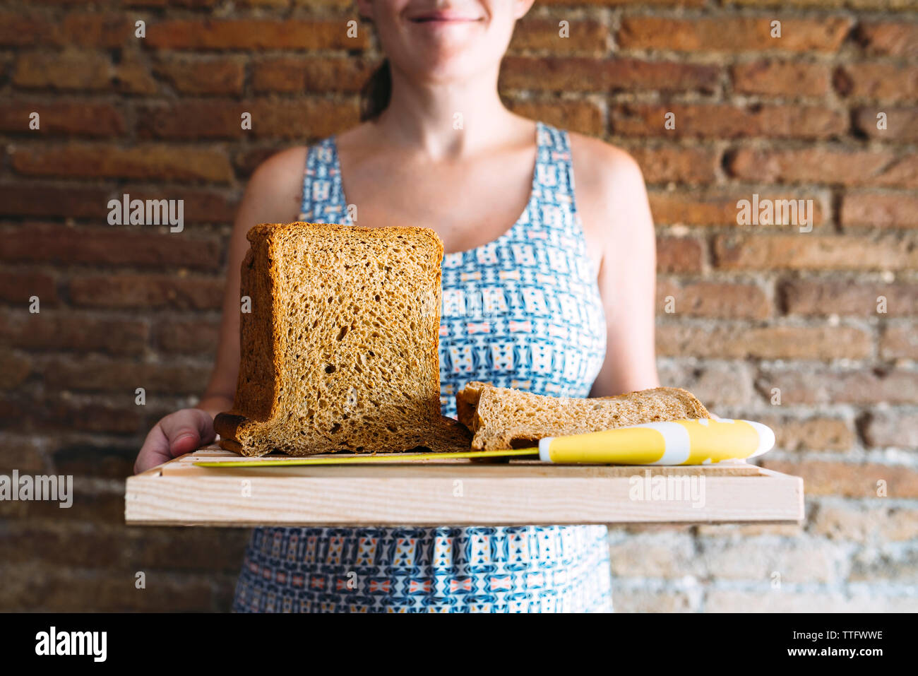 Sección media de la mujer sosteniendo la tabla de cortar con el pan y el cuchillo de cocina contra la pared de ladrillo Foto de stock