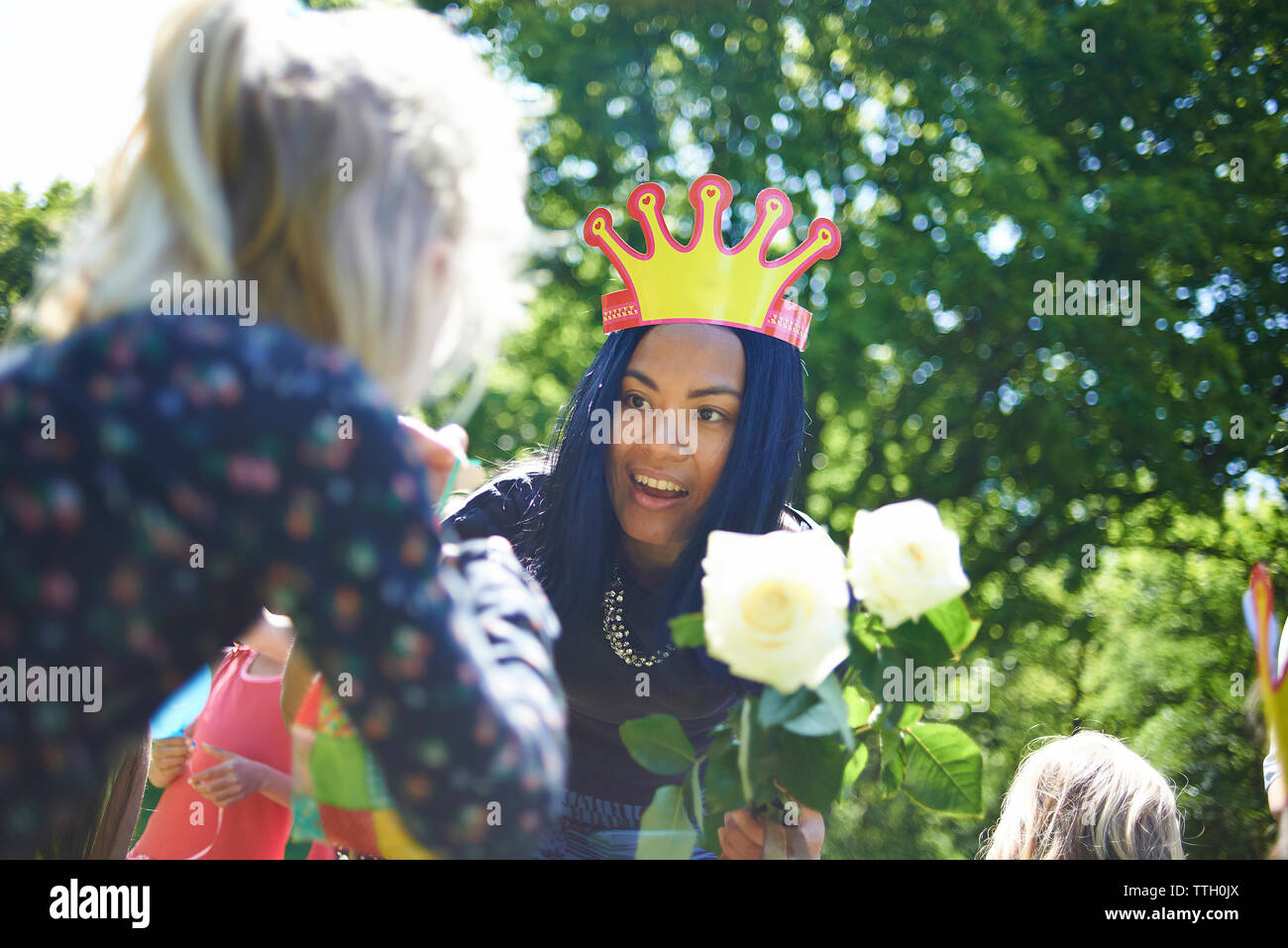 Feliz mujer vistiendo la corona de papel dando rosas blancas al adolescente durante la fiesta de cumpleaños en el patio Foto de stock