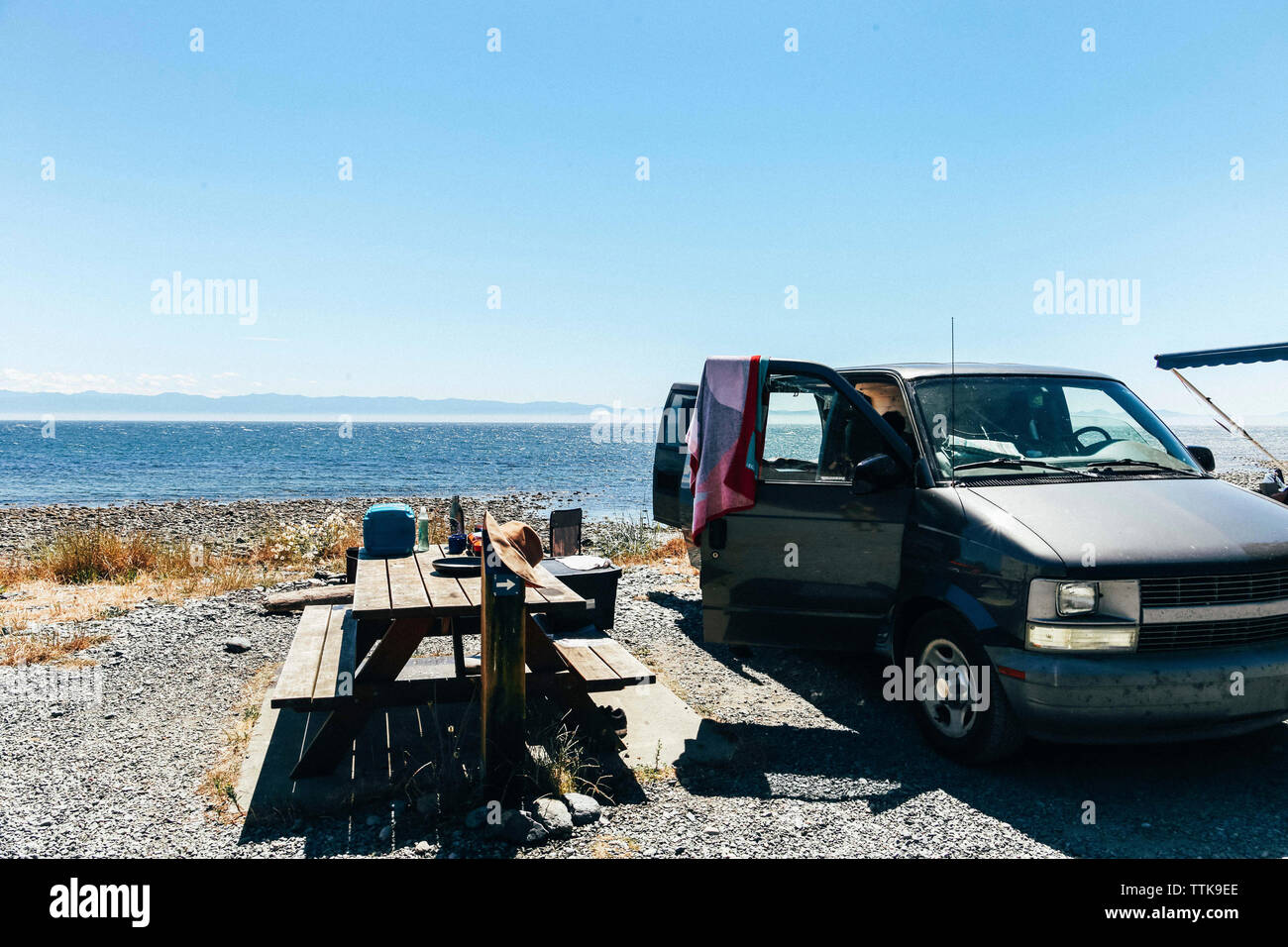 Mesa de picnic y auto abierto en la costa contra el mar y cielo despejado durante el día soleado Foto de stock