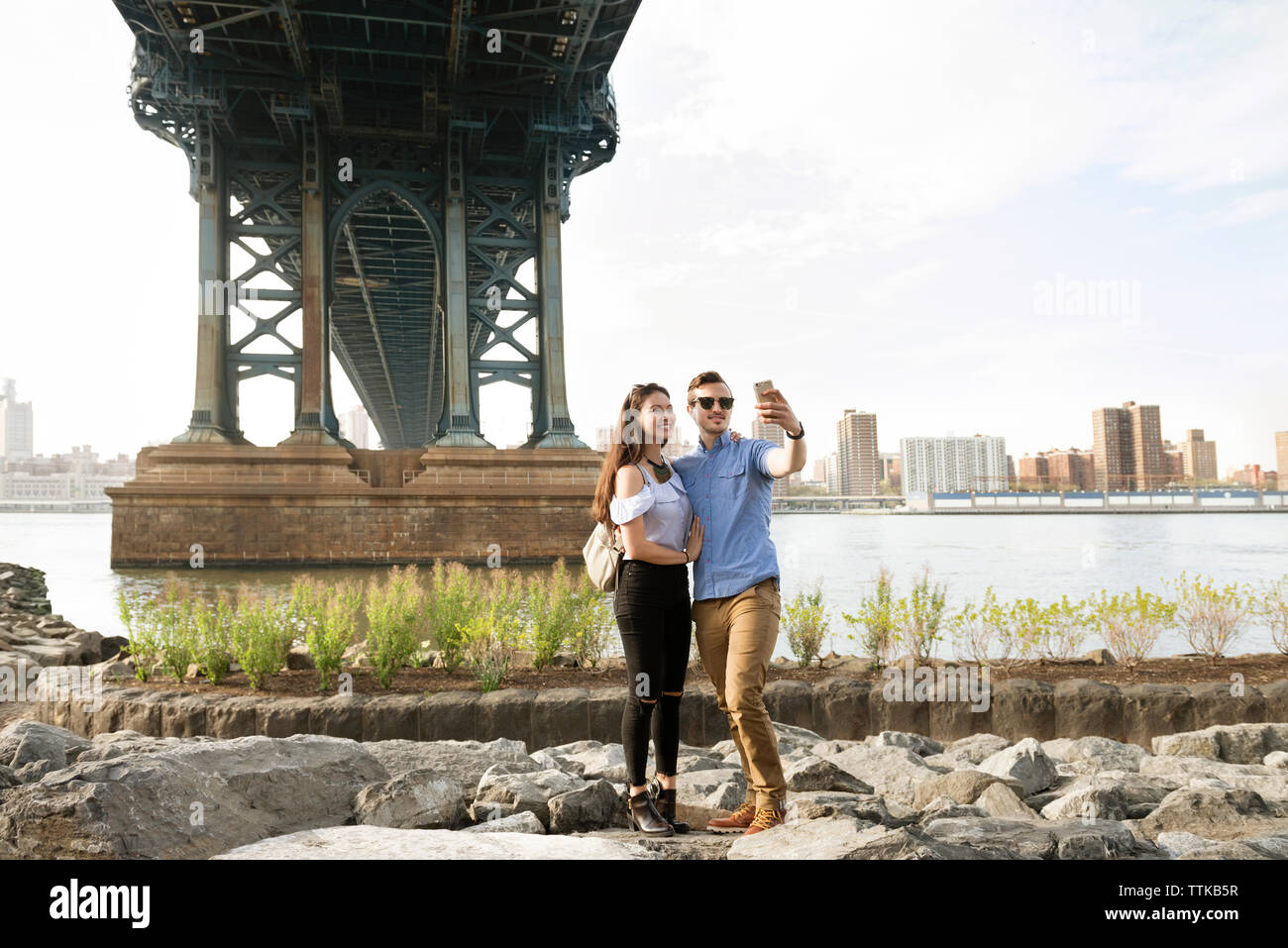 Par tomar selfie estando sobre rocas bajo Manhattan Bridge contra el cielo claro Foto de stock