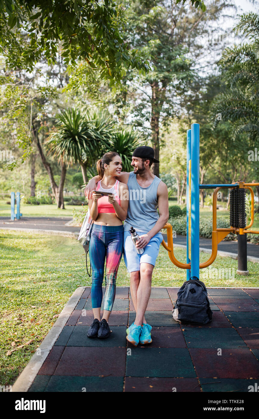 Vista frontal de la feliz pareja recostada sobre equipos de ejercicios en el parque Foto de stock