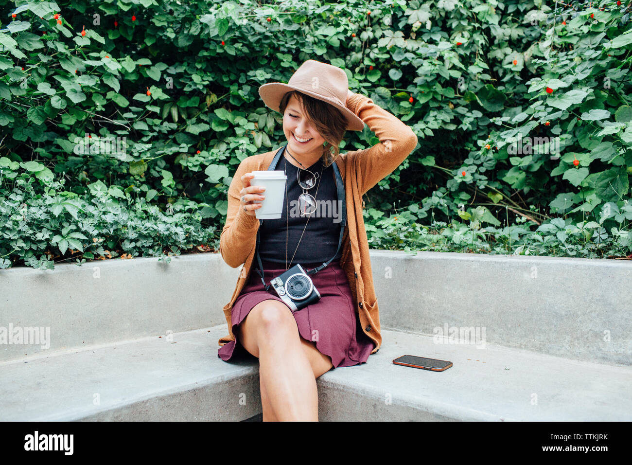 Mujer sonriente Celebración copa desechable mientras está sentado en un banco en el parque Foto de stock