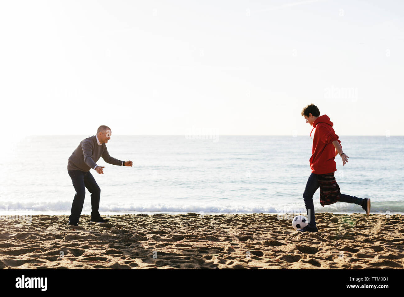 Hijo pateando una pelota de fútbol, mientras que el padre defendiendo a la playa contra el cielo claro Foto de stock