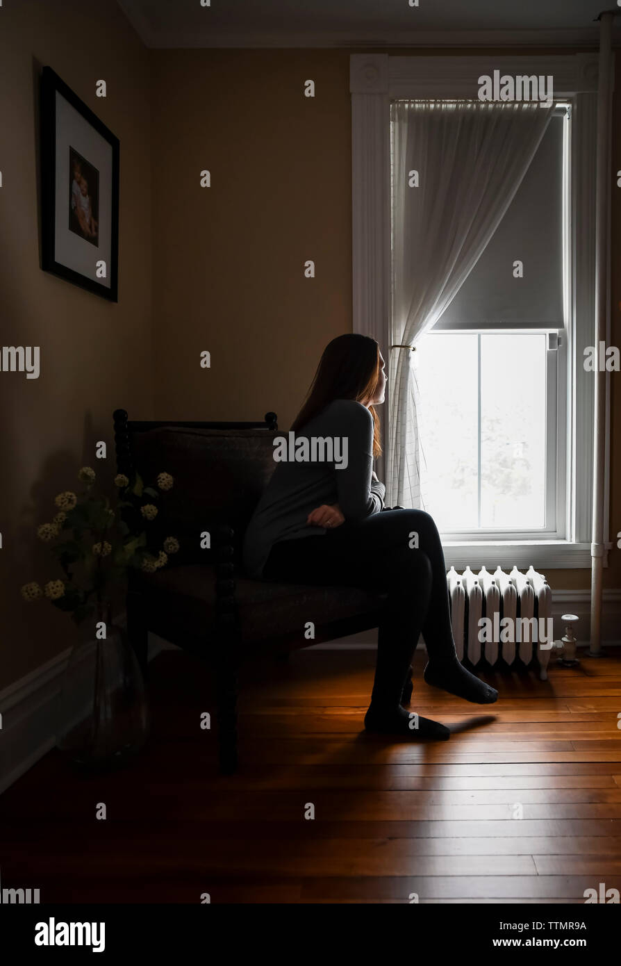 Mujer sentada en una silla en una habitación oscura, mirando hacia afuera de la ventana. Foto de stock