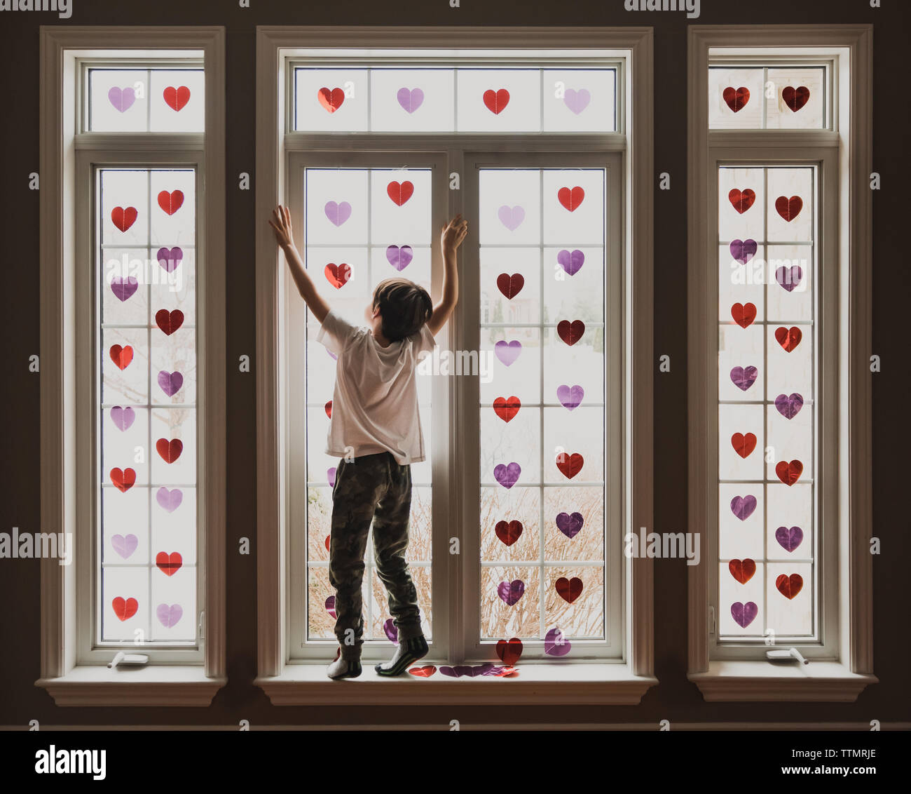 Chico parado en el alféizar de ventanas cubiertas de pequeños corazones brillantes. Foto de stock
