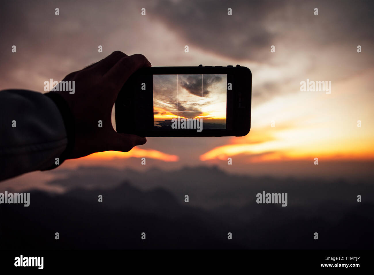 Imagen recortada del hombre fotografiando el cielo nublado a través del teléfono inteligente durante la puesta de sol Foto de stock