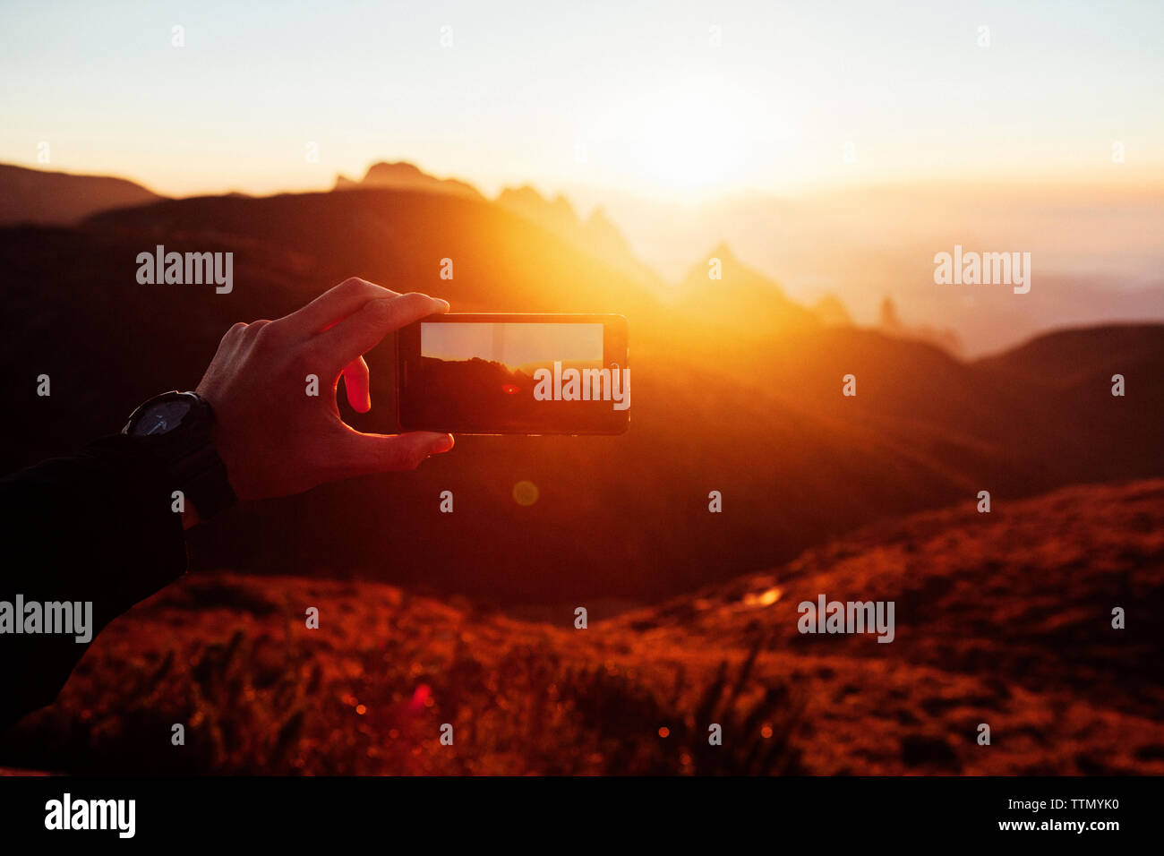 Imagen recortada del hombre fotografiando las montañas a través de teléfono inteligente durante la puesta de sol Foto de stock