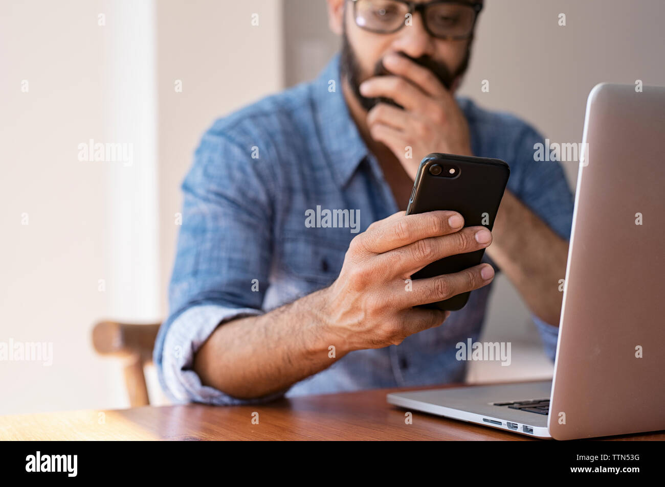 Empresario con ordenador portátil sobre la mesa de madera a través de teléfono móvil mientras está sentada contra la pared en casa Foto de stock
