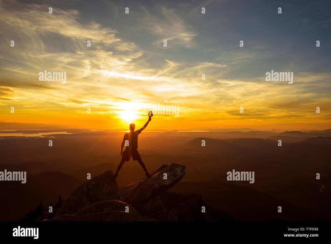 Silueta excursionista sujeta la tapa con la mano levantada mientras está parado en la cima de la montaña durante la puesta de sol Foto de stock