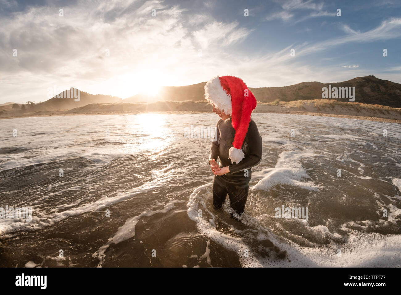 Niño feliz vestidos de rojo Santa hat parado en agua en la playa al anochecer Foto de stock