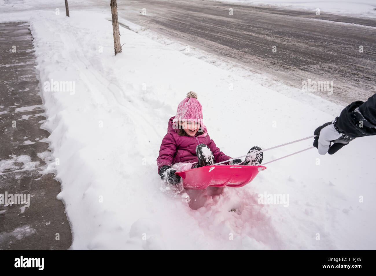 Mano recortada del padre tirando hija sentado en el trineo sobre carreteras cubiertas de nieve Foto de stock