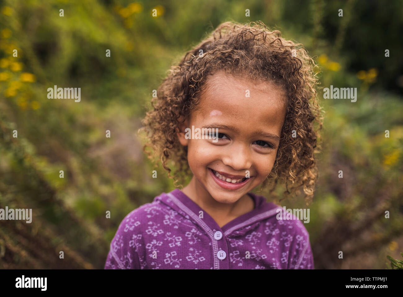 Close-up retrato de niña alegre con el pelo rizado estando en contra de plantas en el parque Foto de stock