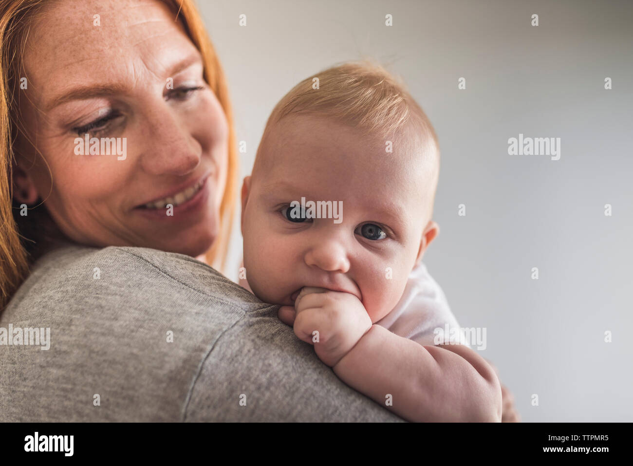 Close-up de madre con hijo lindo con los dedos en la boca mientras está de pie contra la pared gris en casa Foto de stock