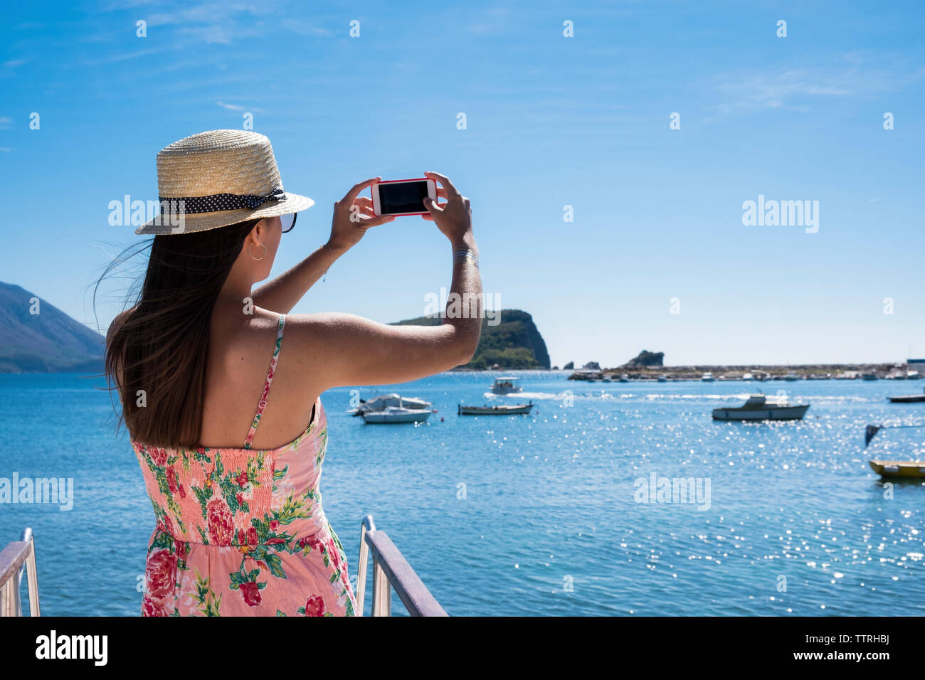 Vista trasera de la mujer fotografiando mar con teléfono inteligente mientras está de pie en el barco contra el cielo azul durante el día soleado Foto de stock