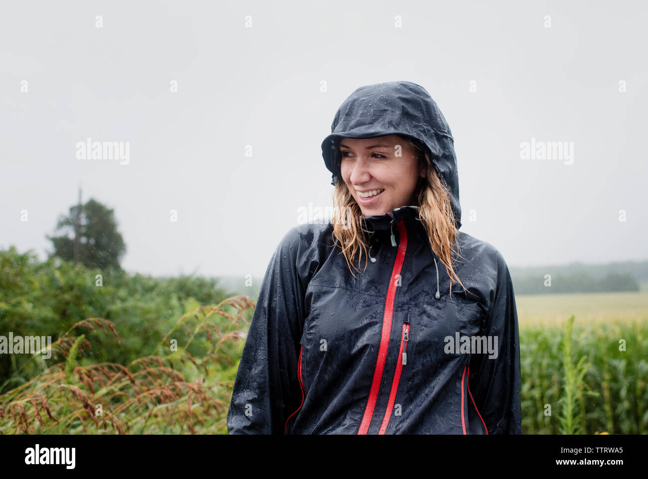 Feliz mujer de impermeable mirando a otro lado mientras está de pie en medio de las plantas contra el cielo durante la temporada de lluvias Foto de stock