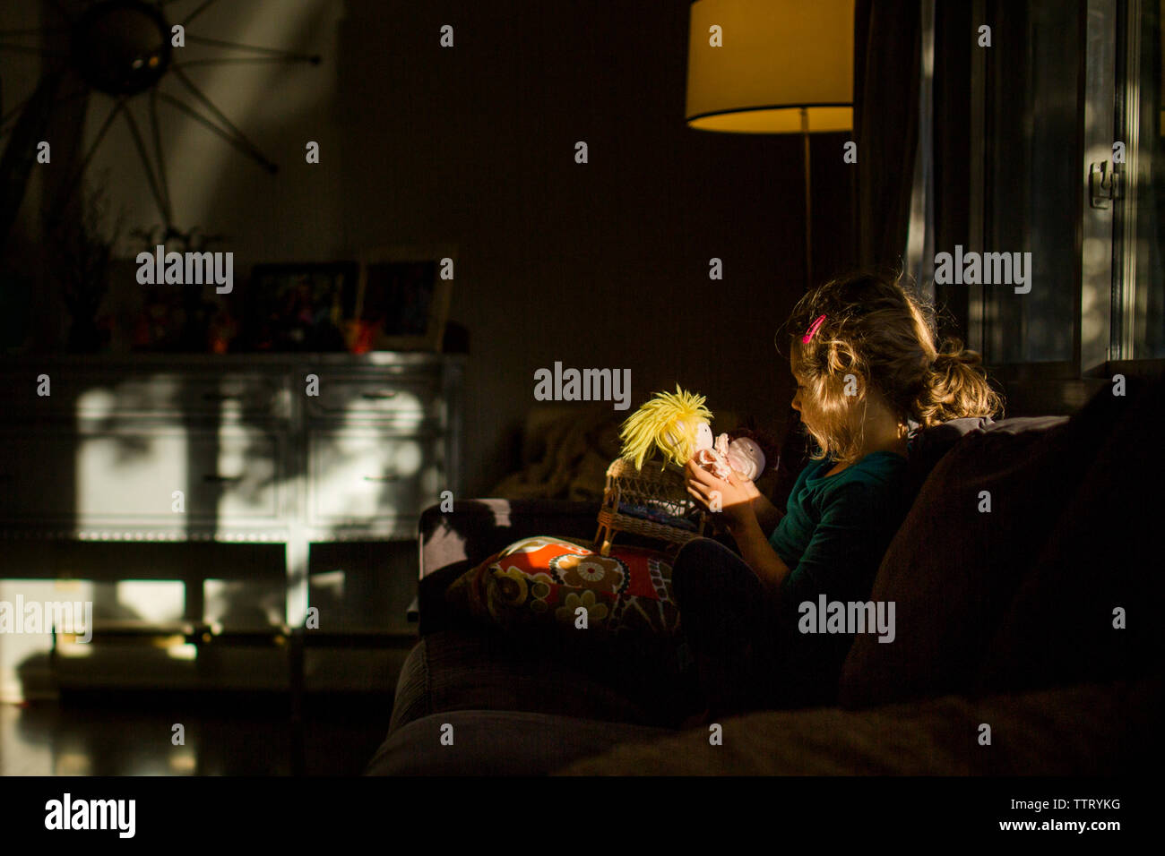 Una niña pequeña está sentada en un parche de luz en una habitación oscura sosteniendo una muñeca Foto de stock