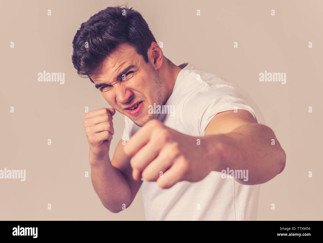 Atractiva joven hombre enfurecido mirando furioso en postura de defensa y amenazan con perforación puño enojado molesto y enojado expresión de cara gris aislado ba Foto de stock