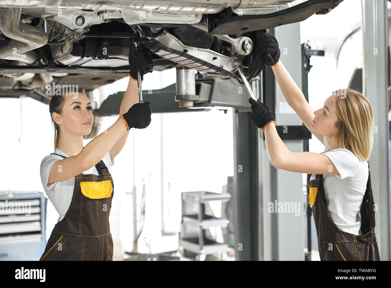 Dos mujeres pararse bajo el coche de mecánica y taller de reparación de  chasis del auto. Mujeres fuertes en monos y guantes protectores trabajando  en el Salón del Automóvil de fijación y