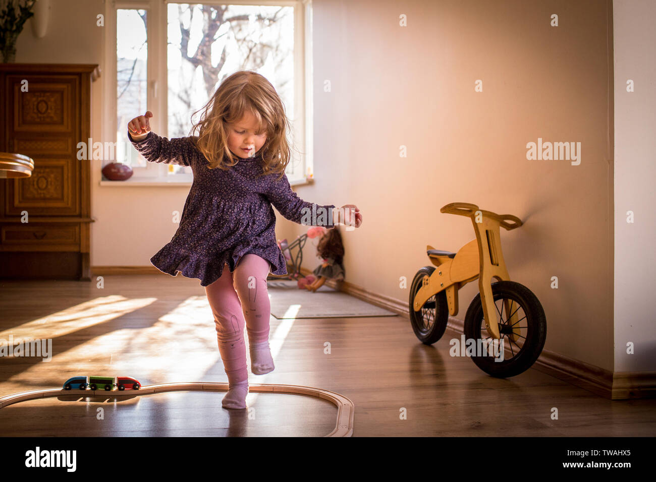 Feliz, pequeño niño niña jugando y bailando dentro de las vías de tren de juguete, en una soleada habitación con ventana, divertirse durante su repr. Foto de stock