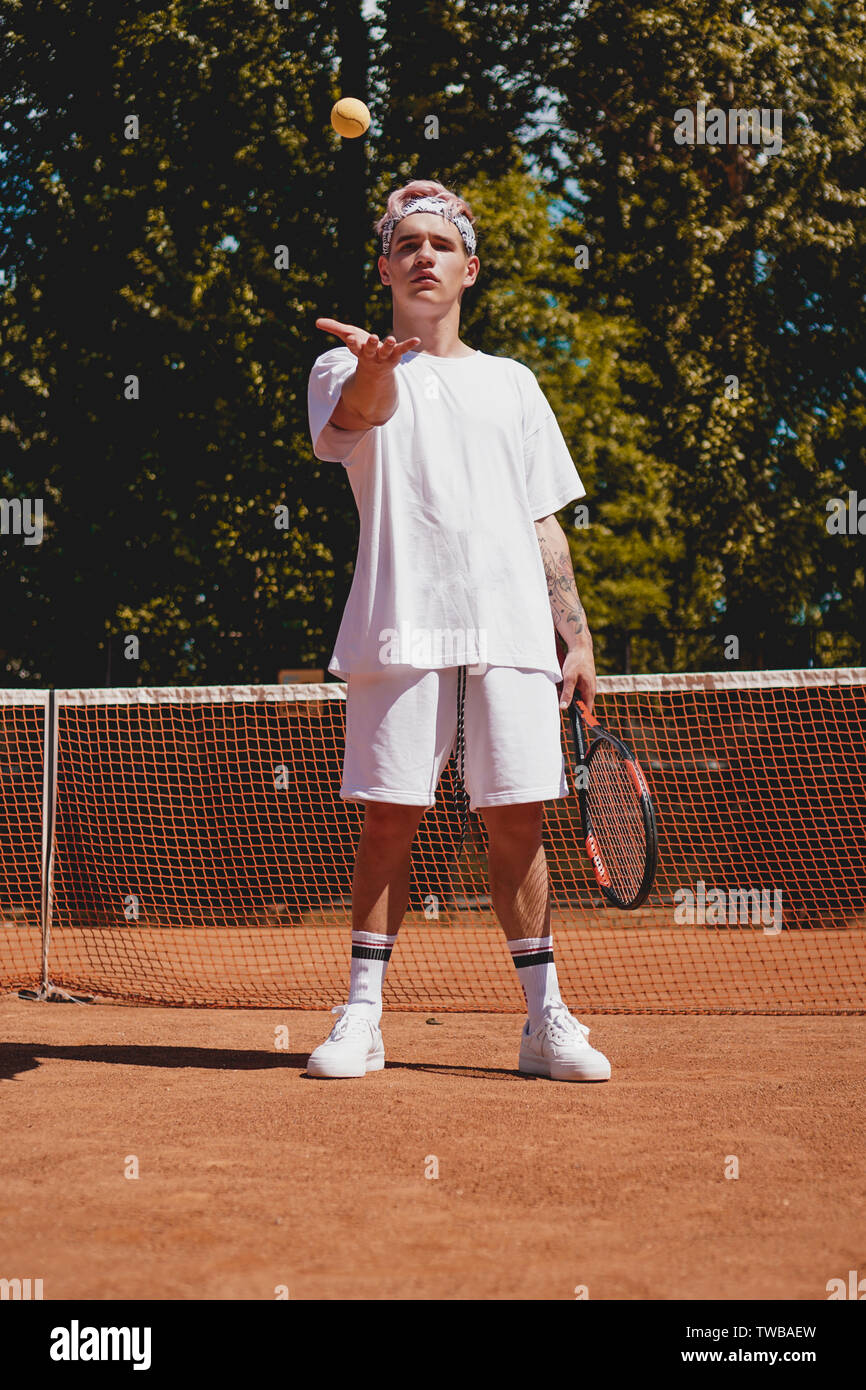 El deporte del tenis. Hombre jugando al tenis al aire libre. Retrato de  joven atractivo en camiseta blanca con la raqueta y la bola en el fondo del  cielo. Un estilo de