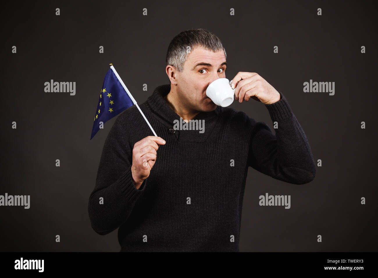 Hombre sujetando la bandera de EU. Hombre en un suéter oscuro sobre un  fondo negro posee la bandera de la Unión Europea y una taza de café y  bebidas Fotografía de stock -