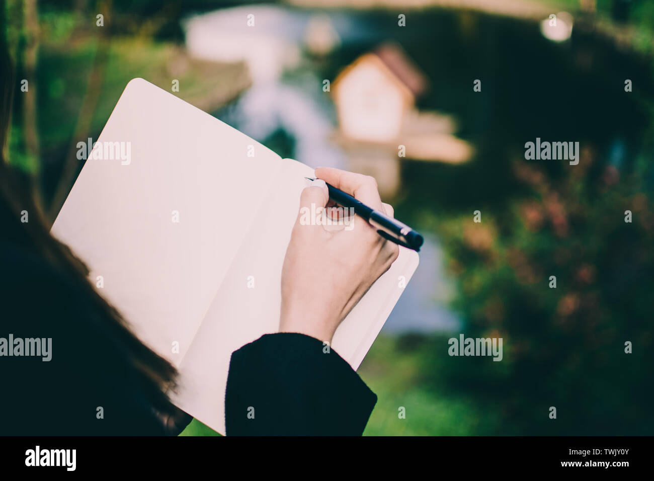 Chica escribiendo sus pensamientos en un cuaderno contra el pequeño cisne borrosa casa en medio de un estanque en un parque. Mano pálida y colorido telón de fondo. Foto de stock