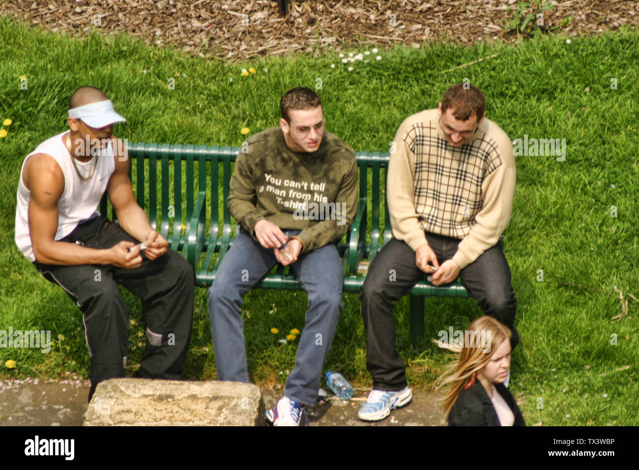 Tres jóvenes que miran a una chica pasando mientras preparan una junta sentada en un banco en un parque público, al sur de Londres, Inglaterra, Gran Bretaña, Reino Unido. Foto de stock