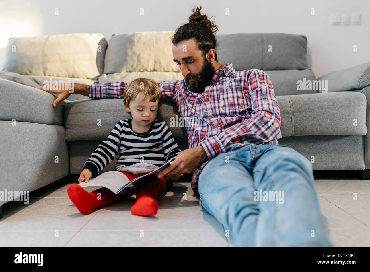 Padre e hija sentado en el piso en el salón leyendo un libro juntos Foto de stock