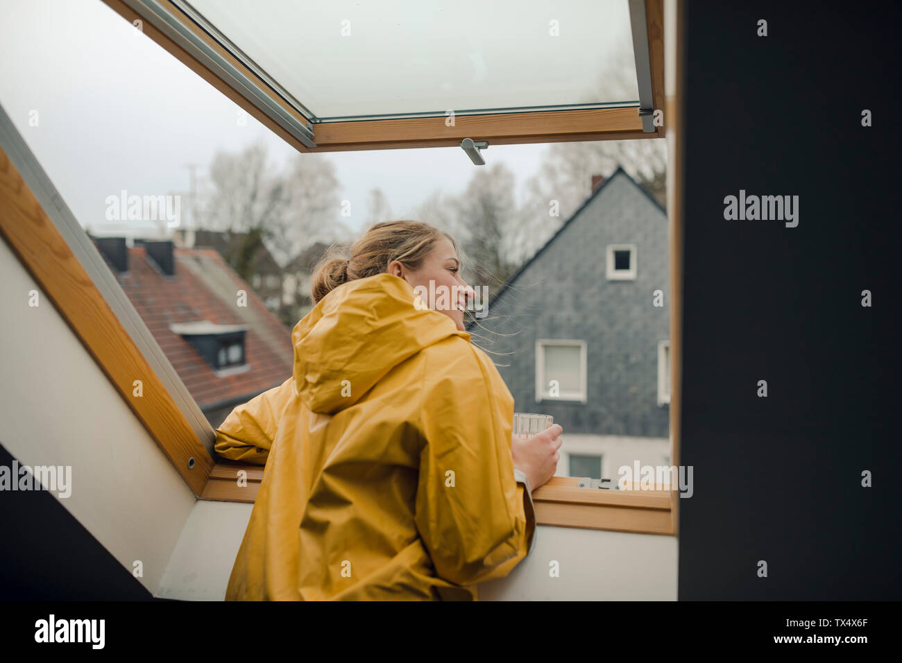 Mujer sonriente en impermeable mirando por la ventana el ático Foto de stock