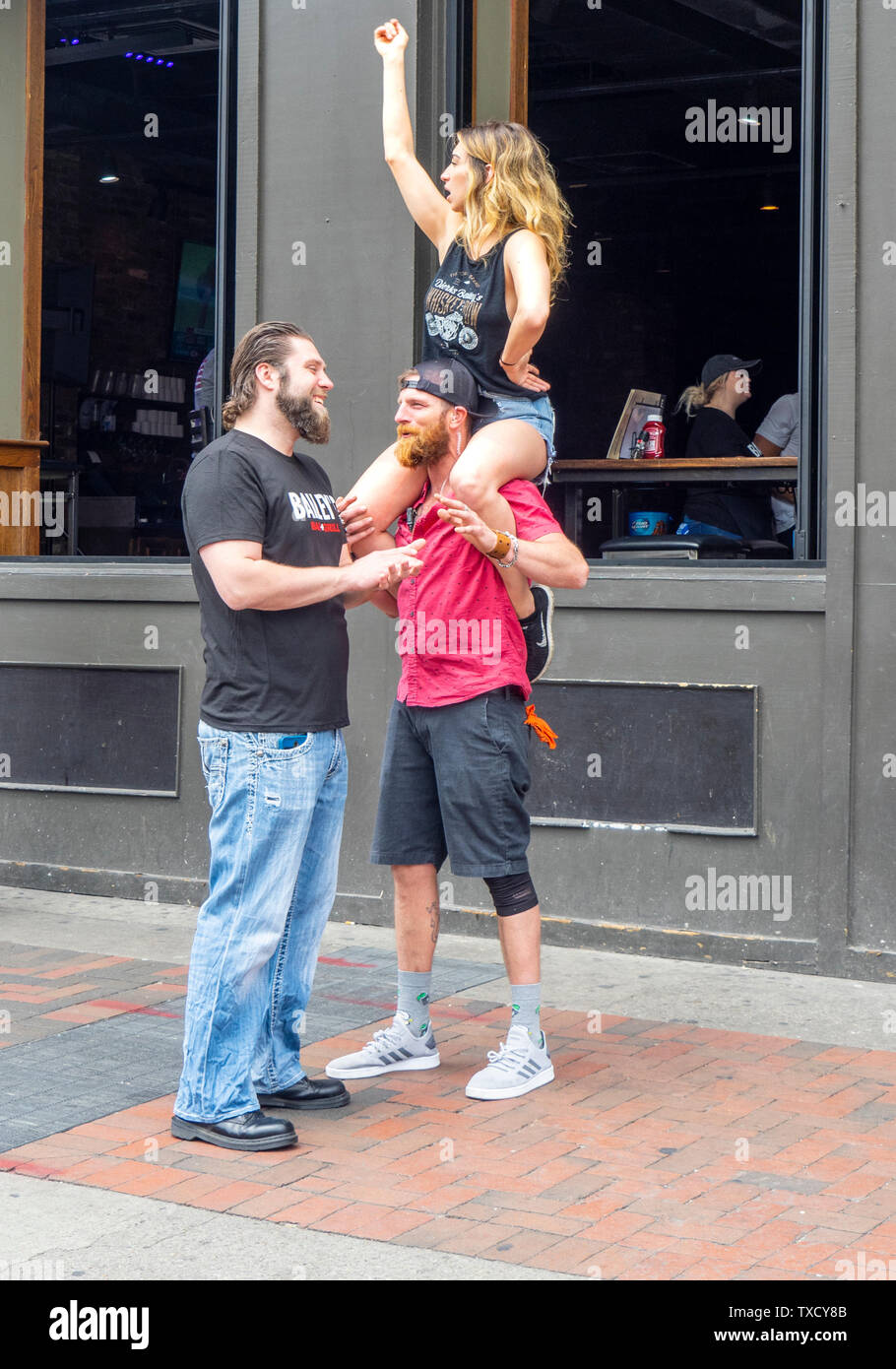 Una mujer sentada sobre los hombros del hombre, mientras él está hablando con otro hombre en Broadway en el NFL Draft 2019 de Nashville, Tennessee, EE.UU.. Foto de stock