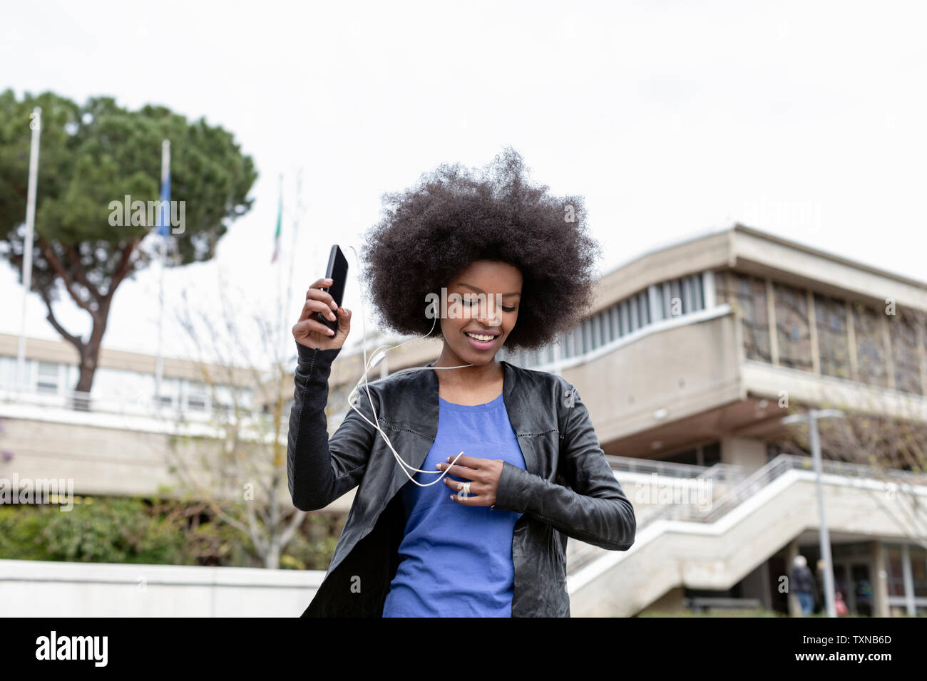 Mujer joven con cabello afro en la ciudad, caminando y escuchando música del smartphone Foto de stock