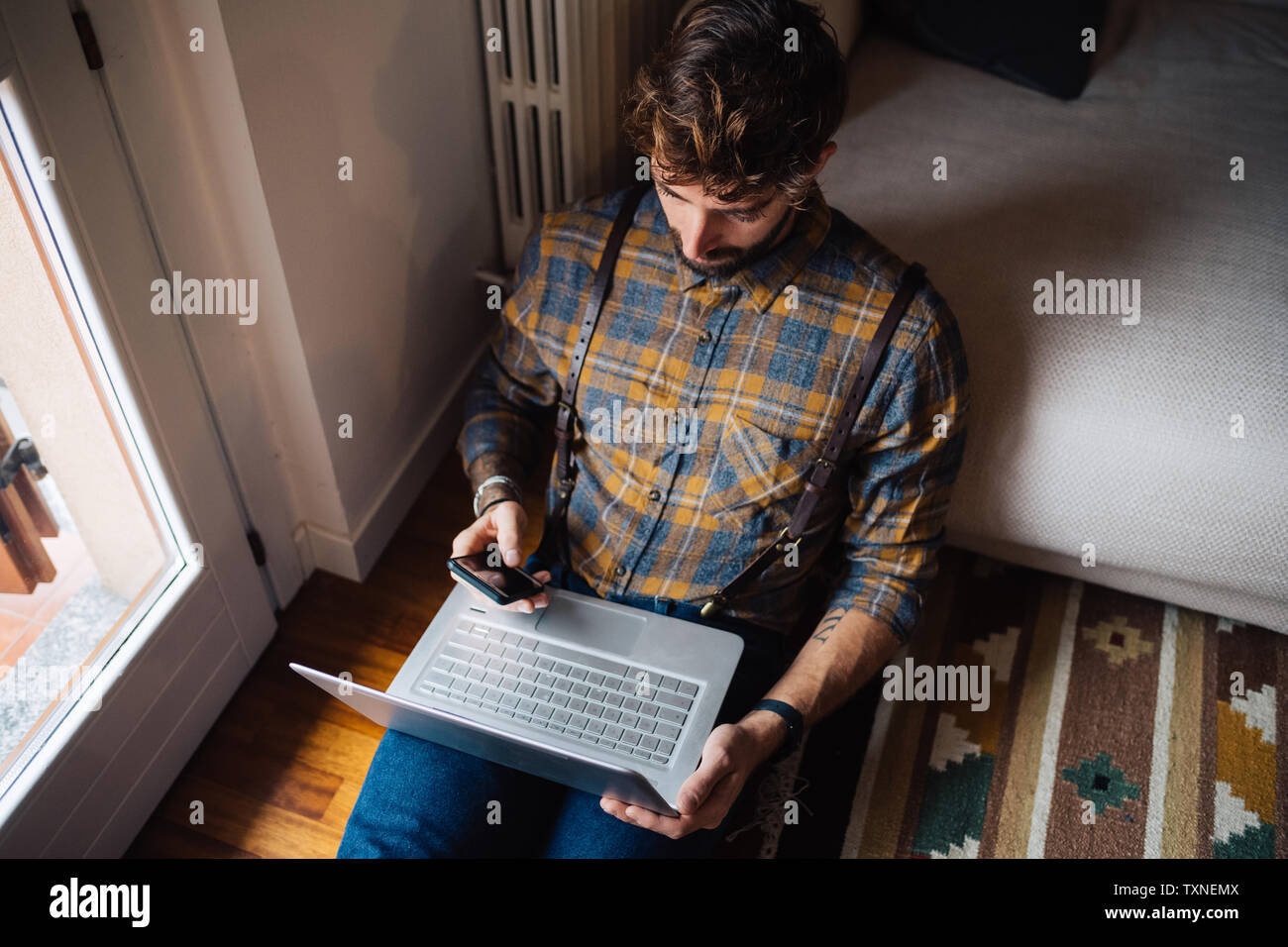 Mitad hombre adulto sentado en el suelo del cuarto de estar con ordenador portátil utilizando el smartphone Foto de stock