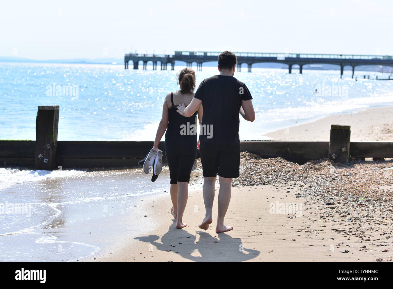 Pareja joven romántica caminando en la playa en verano con los zapatos quitados y tocando cariñosamente, Boscombe, Bournemouth, Dorset, Inglaterra, Reino Unido, junio de 2019 Foto de stock