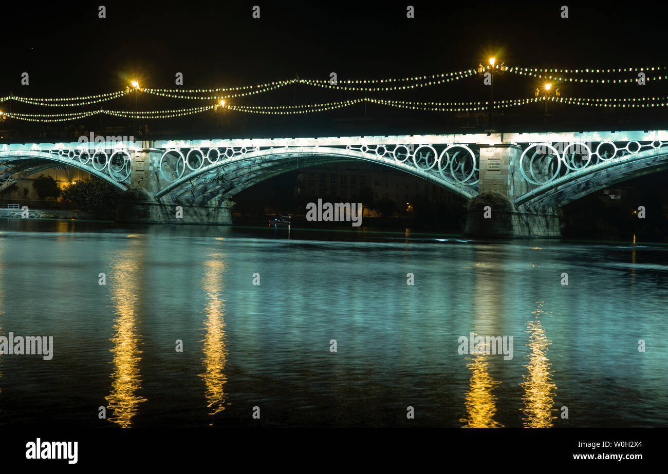 El río Guadalquivir a medida que pasa por el Puente de Triana iluminado en la noche, Sevilla Foto de stock