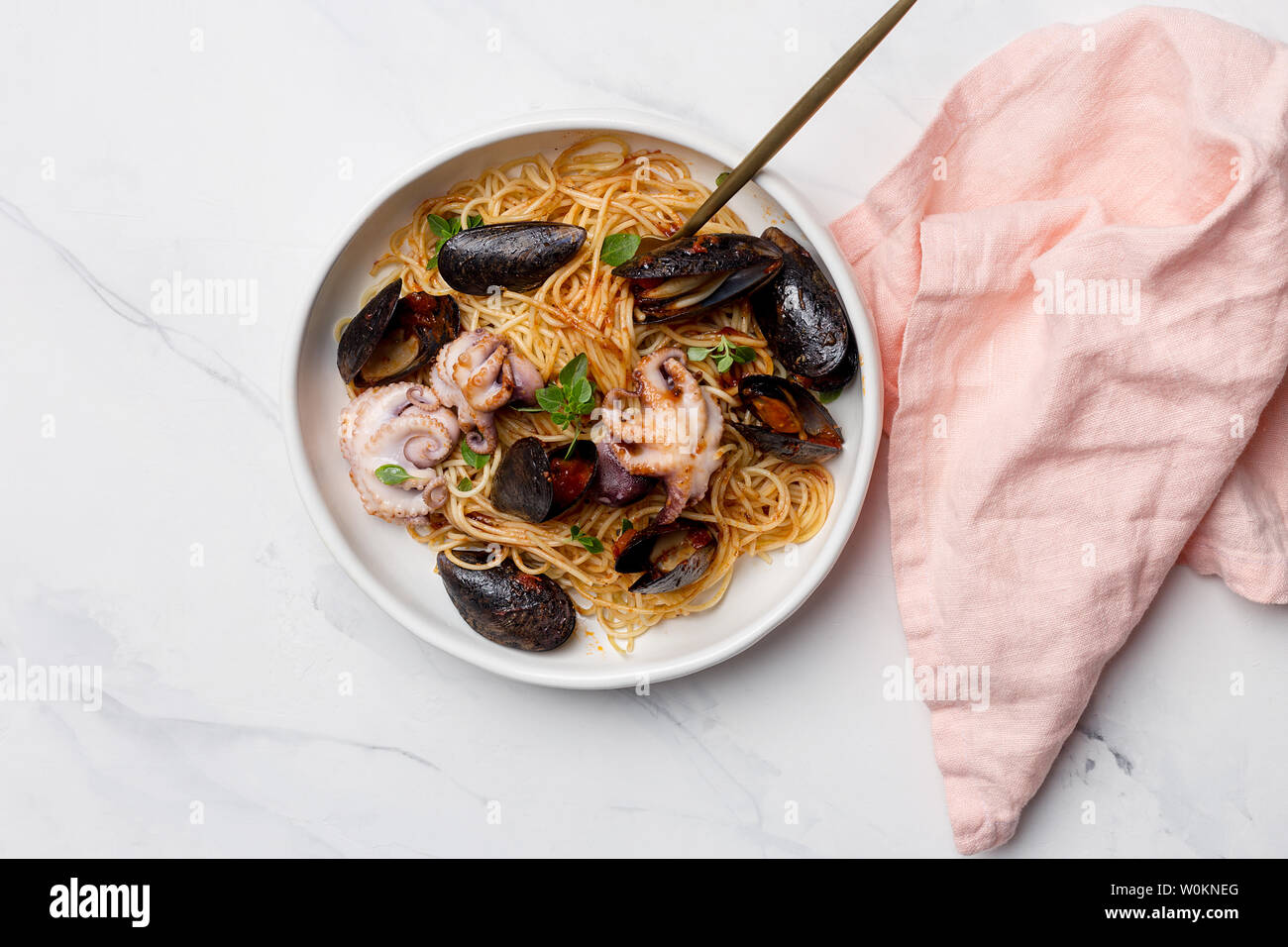 Pasta con marisco y salsa de tomate. Espaguetis con pulpo y mejillones en tazón blanco con un tenedor y servilleta rosa sobre fondo blanco. Concepto de seafoo Foto de stock