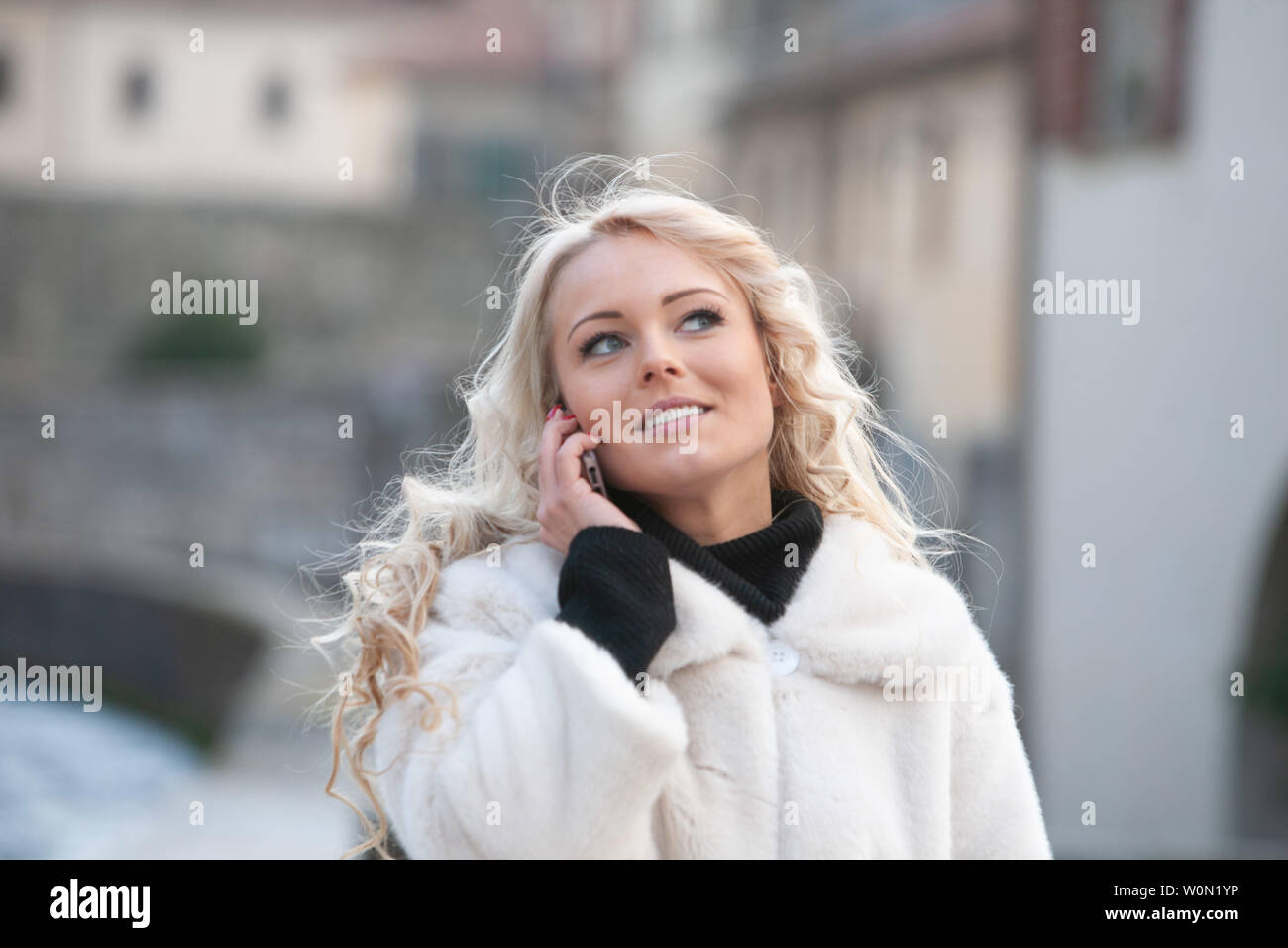 Joven elegante mujer rubia en un abrigo blanco caminando por la ciudad mediante un móvil escuchando la conversación con una amable sonrisa Foto de stock