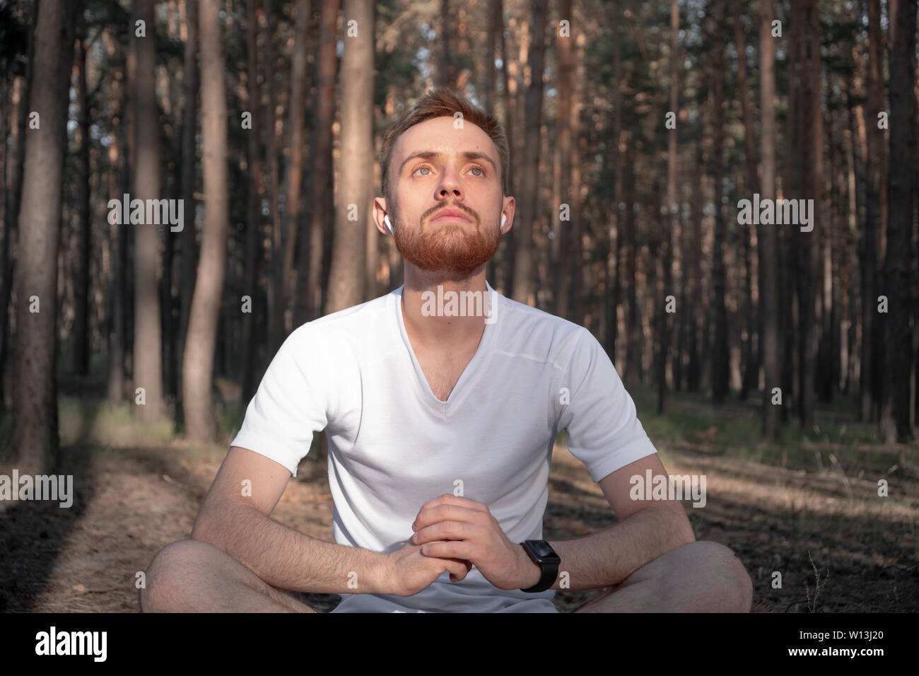 Macho joven persona meditando en el bosque, utilizando tecnología moderna. El hombre se sienta activo en los bosques de pinos y goza de la meditación al aire libre Foto de stock