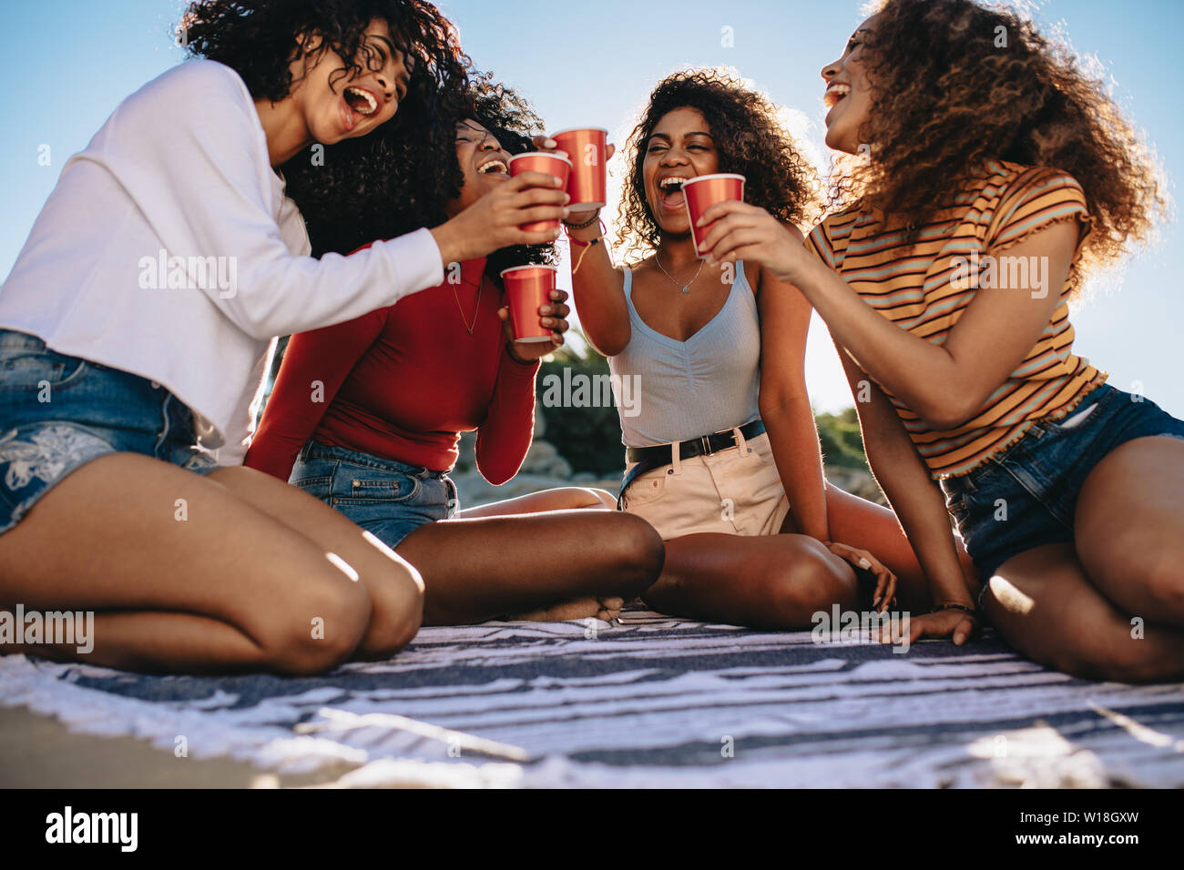 Grupo de mujeres sentados juntos en Manta y bebidas de tostado. Las mujeres alegres amigos haber cola en la playa. Foto de stock