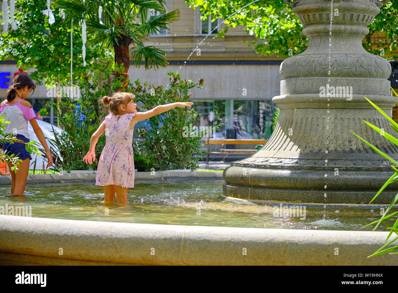 Muchacha con un vestido de sol jugando en el parque la fuente. De pie en las rodillas agua, levantando la mano para recoger el goteo hot día soleado en Zagreb Foto de stock