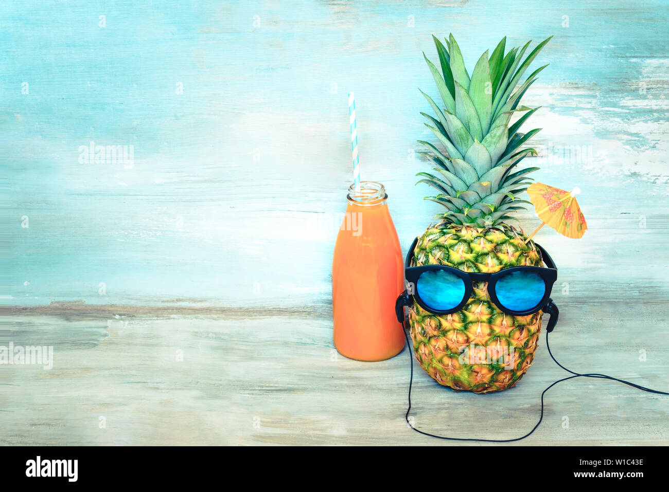 Piña con gafas de sol y auriculares y una botella de jugo en frente de un azul de fondo rústico de madera. Concepto de fiesta de vacaciones de verano. Foto de stock