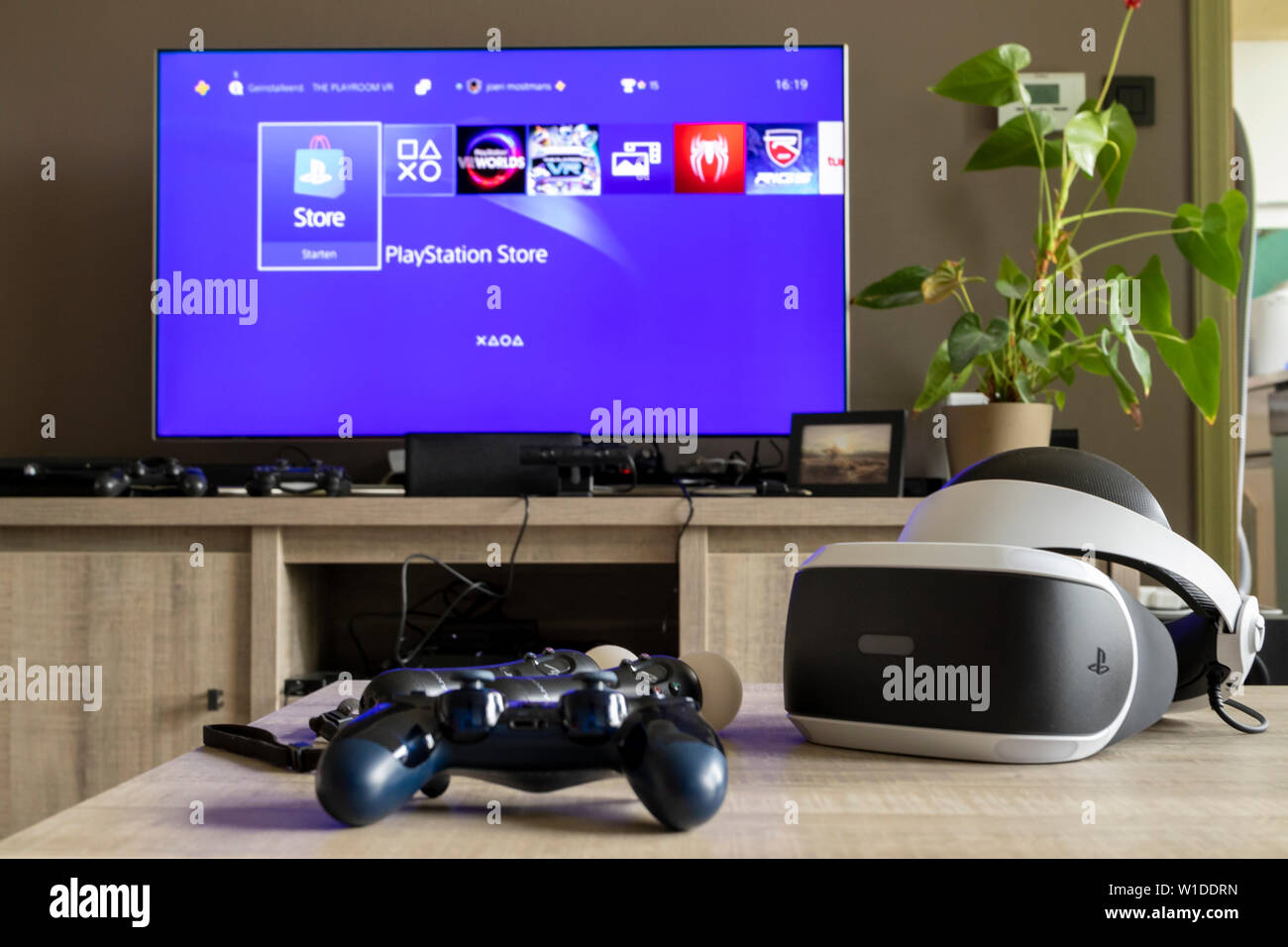 Un auricular VR de playstation en una mesa de madera junto con un  controlador de PS4 y algunos controladores de playstation move. En el fondo  hay un televisor Fotografía de stock -