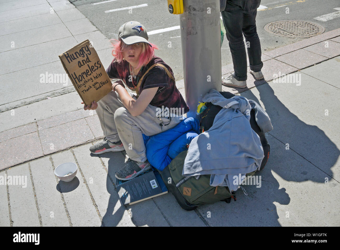 Homeless chica con pelo de color rosa claro ojos azules sombreadas por una gorra de béisbol se asienta en bedroll en la base farola aguantando miserable firmar en el centro de Ottawa Foto de stock
