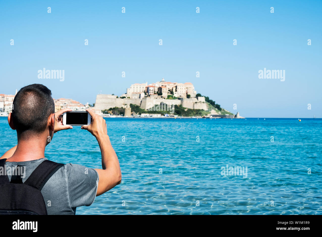 Un joven hombre caucásico, visto desde atrás, tomar una foto de Calvi, en Córcega, Francia, con el mar Mediterráneo en primer plano y la ciudadela o Foto de stock