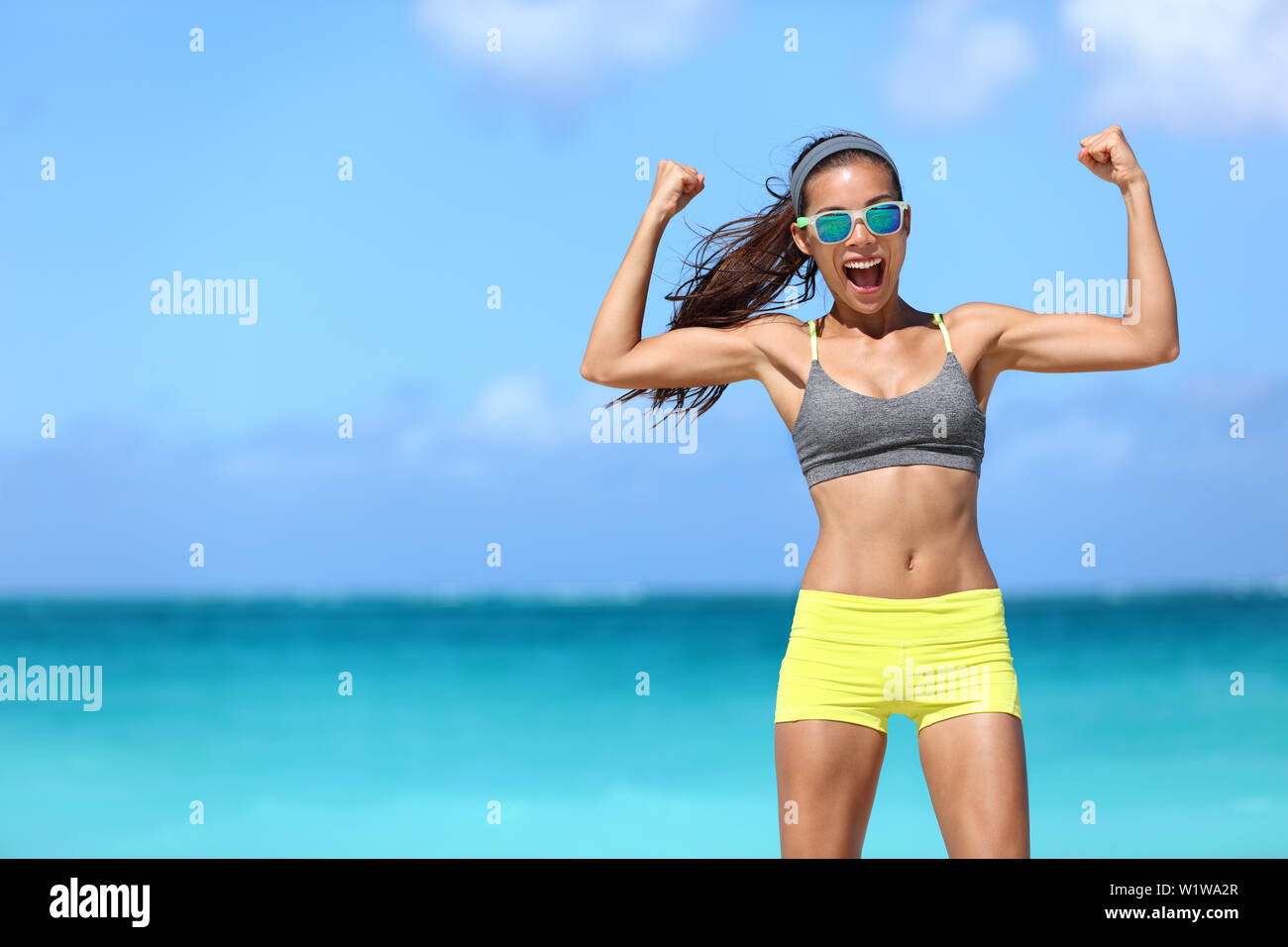 Fuerte fitness mujer divertida en neón azul caminante gafas de sol en la playa mostrando muscular bíceps flexión de los brazos para la diversión. Colocar chica en ropa deportiva tras la ejecución de ejercicios de entrenamiento de fuerza ganando en potencia. Foto de stock