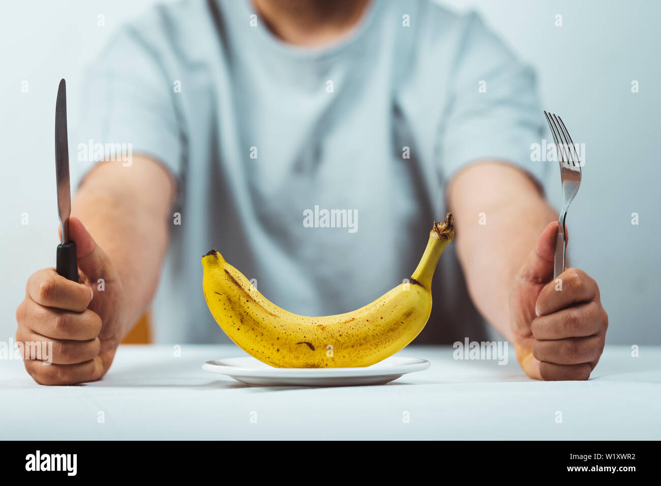 Hombre sentado detrás de una mesa con un tenedor y un cuchillo en la mano y un plátano fresco sobre una placa en una tabla blanca , la hora de comer - Alimentación saludable concepto Foto de stock