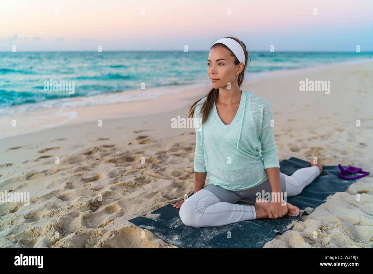 Esterilla de Yoga mujer estirando los músculos de la cadera, el muslo y la ingle, los músculos de la pierna con la paloma plantean stretch. Colocar asiática chica atleta fitness deportes ejercicio estiramientos activewear en la playa al atardecer. Foto de stock