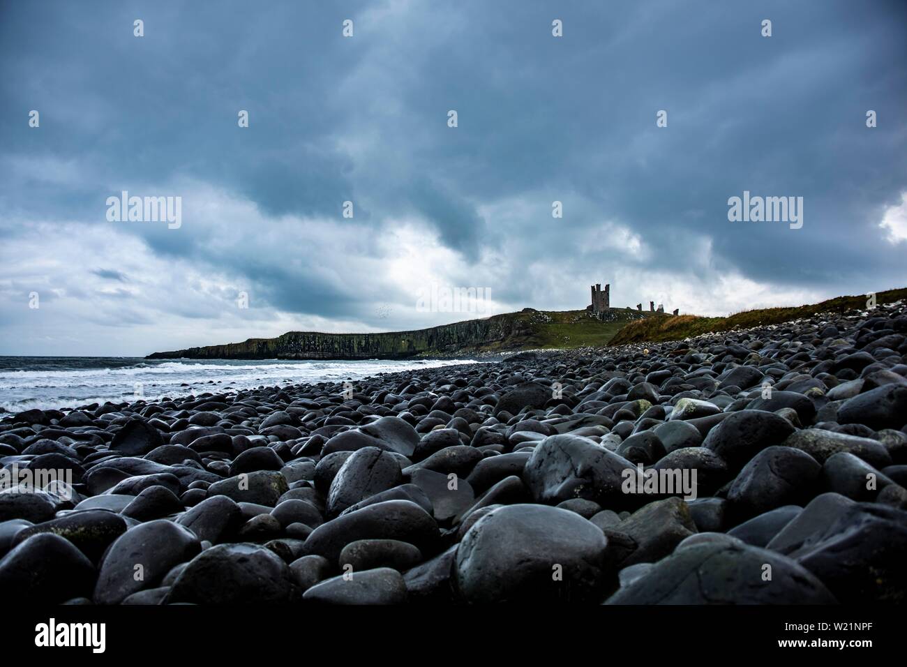 Negro, ronda de las rocas de la costa, el castillo de Dunstanburgh con cielo nublado en la espalda, Craster, Northumberland, Gran Bretaña Foto de stock