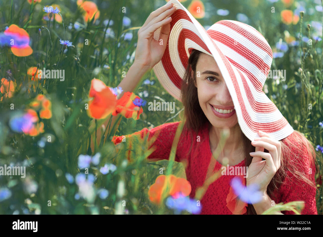 Mujer de ensueño en el vestido rojo y un gran sombrero de rayas rojas está sentado en la hermosa hierba floración verano campo de amapolas. Vintage elegante aspecto romántico concepto. Foto de stock