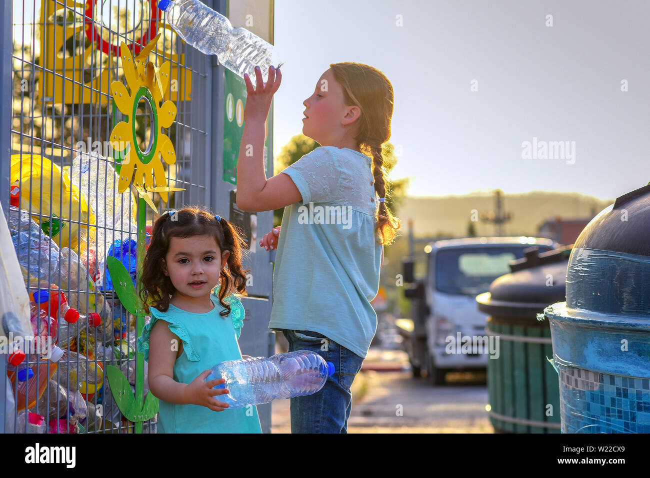 Las niñas de plástico reciclado de botellas de agua en la jaula de Reciclaje de metal amarillo. Los niños pone residuos plásticos en contenedores de reciclaje en la calle de la ciudad. Foto de stock